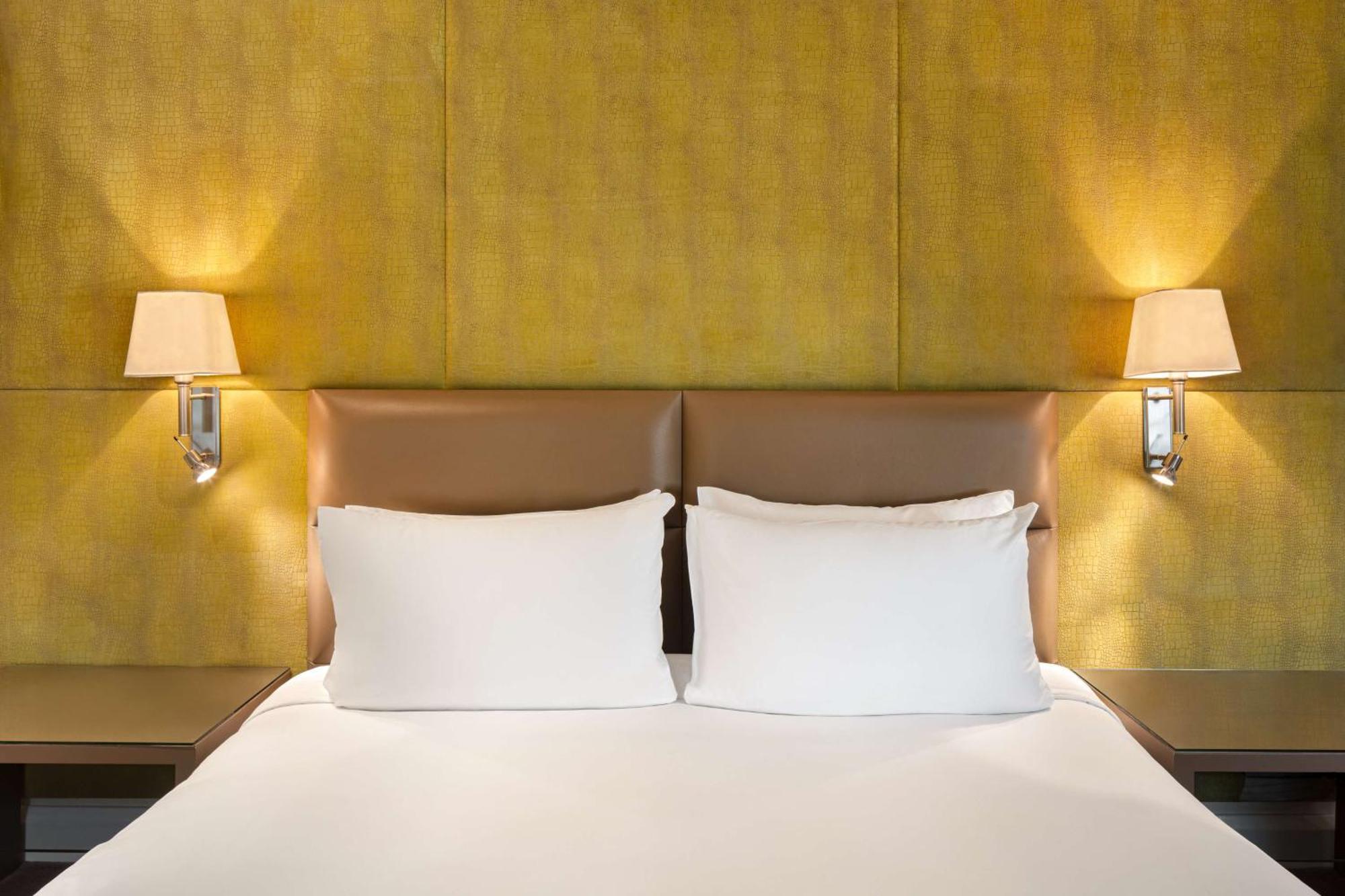 The May Fair, A Radisson Collection Hotel, Mayfair Лондон Экстерьер фото The photo shows a cozy bedroom setting featuring a large bed with two white pillows. The headboard has a smooth, light brown leather material, and there are two wall-mounted lamps on either side of the bed, illuminating a textured yellow wall behind 