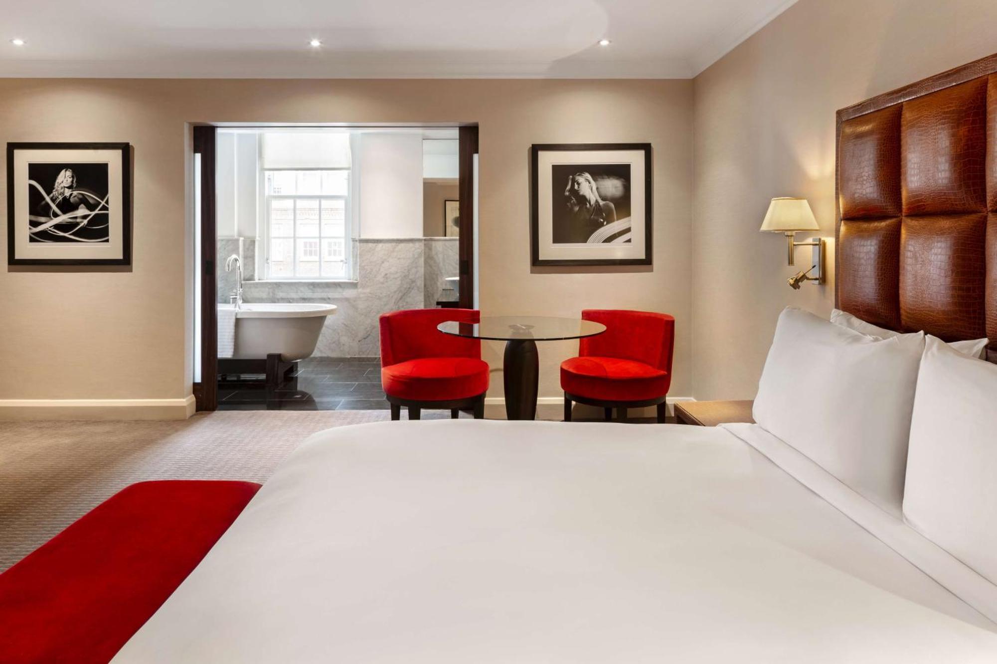 The May Fair, A Radisson Collection Hotel, Mayfair Лондон Экстерьер фото The photo shows a hotel room featuring a large bed with white linens and a red accent throw. In the background, there are two modern red chairs around a small glass table. The wall displays framed black and white photographs. To the side, there is an